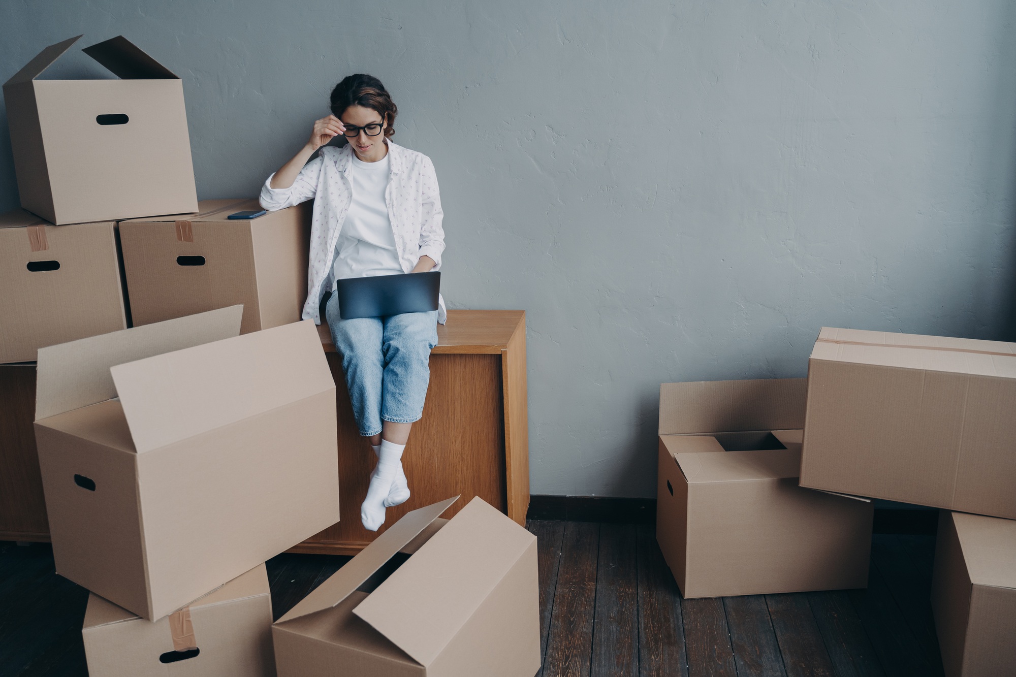 Businesswoman working at laptop sitting surrounded by cardboard boxes for moving in new office